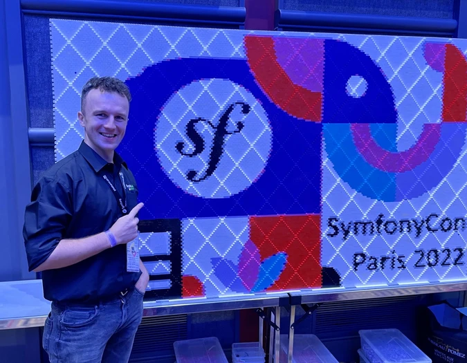 Mann posiert neben einer SymfonyCon Paris 2022 Pixel-Art-Wand