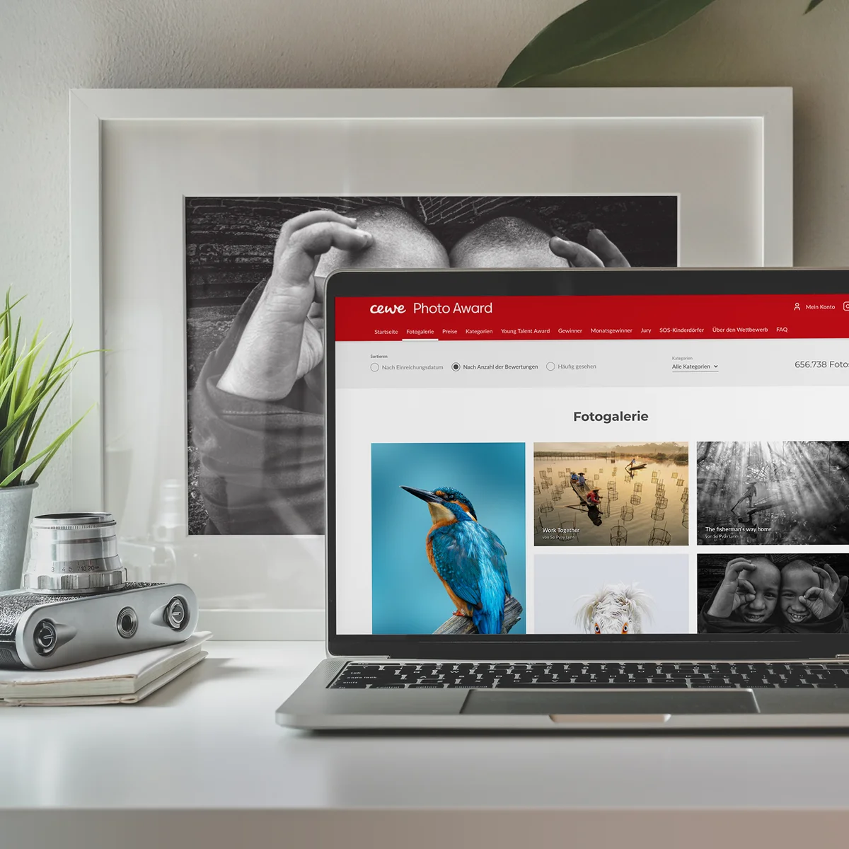 CEWE E-Commerce-Website-Vorschau auf einem Laptop mit der Galerie des „CEWE Photo Award“ und verschiedenen preisgekrönten Fotografien vor einem Schreibtisch-Hintergrund mit einem gerahmten Schwarz-Weiß-Foto und einer Vintage-Kamera