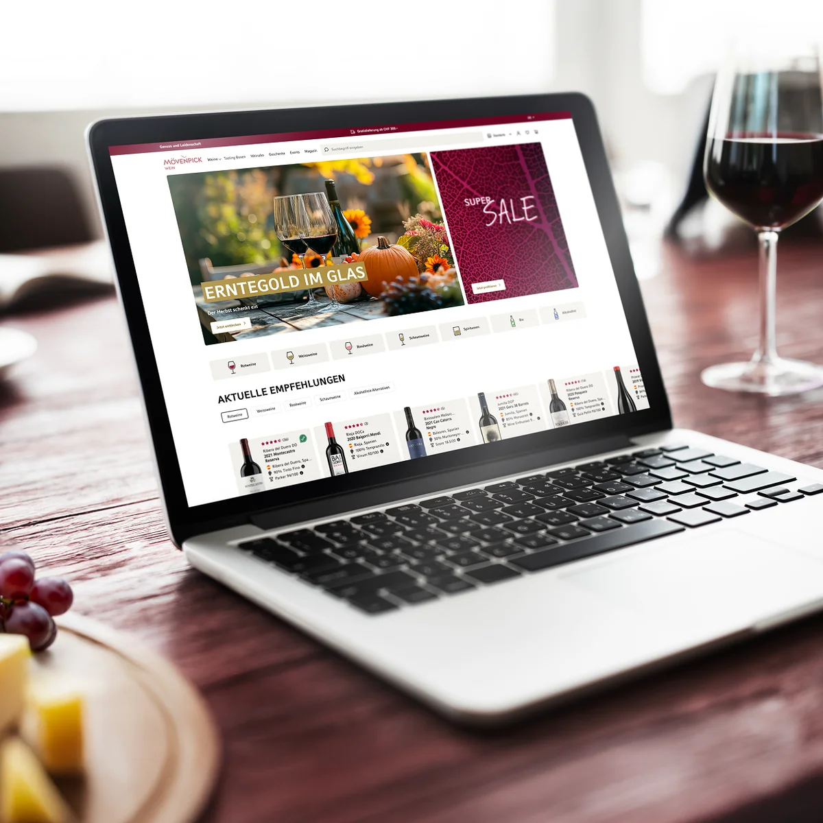 Mövenpick Wein E-Commerce-Website-Vorschau auf einem Laptop mit herbstlichen Promotionen und aktuellen Weinempfehlungen vor einem stimmungsvollen Gastronomie-Hintergrund mit einem Glas Rotwein, Weintrauben und Käse
