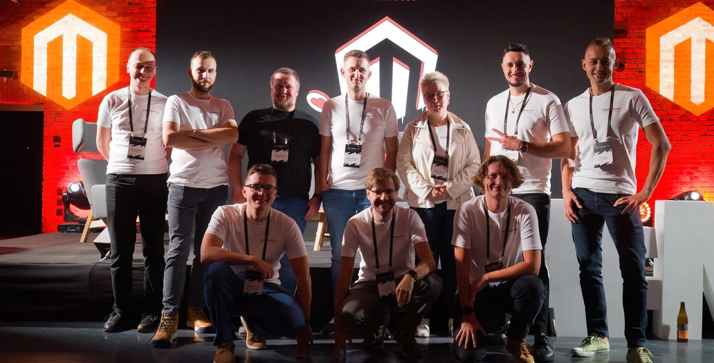 Eine Gruppe von Teammitgliedern, darunter CEO Jaromir, posiert auf einer Bühne beim Meet Magento PL Poznań vor einem großen beleuchteten Logo