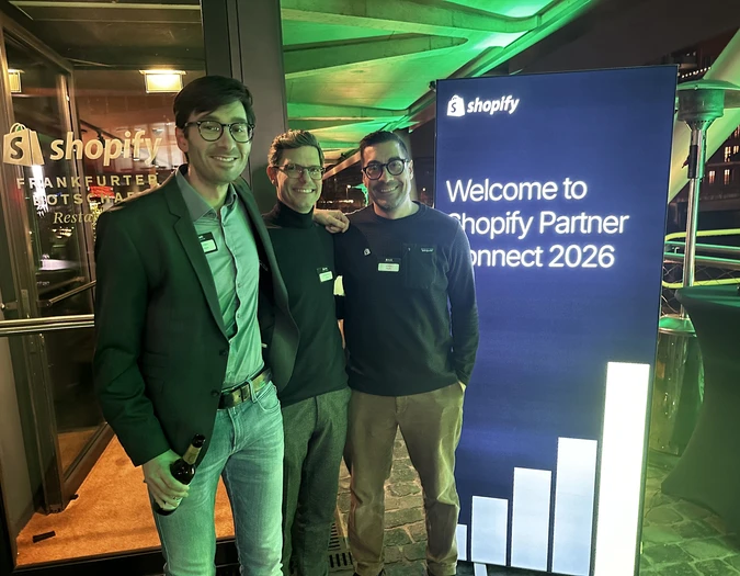 Drei Männer posieren am Eingang eines Shopify-Events neben einem blauen „Welcome to Shopify Partner Connect 2026“ Schild