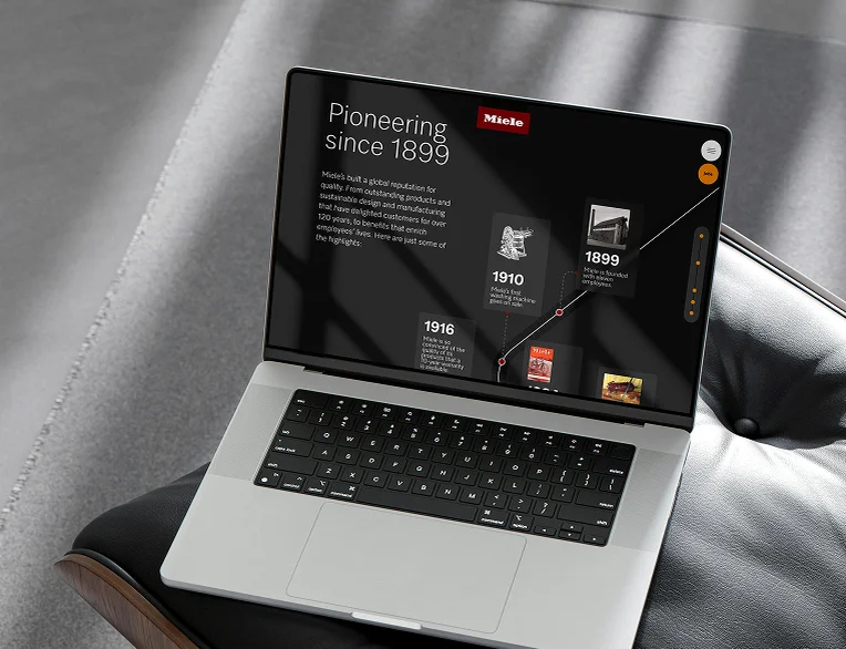 Miele X E-Commerce-Website-Vorschau auf einem Laptop mit einer Markenhistorie-Zeitleiste „Pioneering since 1899“, die Meilensteine wie die erste Waschmaschine von 1910 zeigt, vor einem dunklen Hintergrund in einem hochwertigen Interieur
