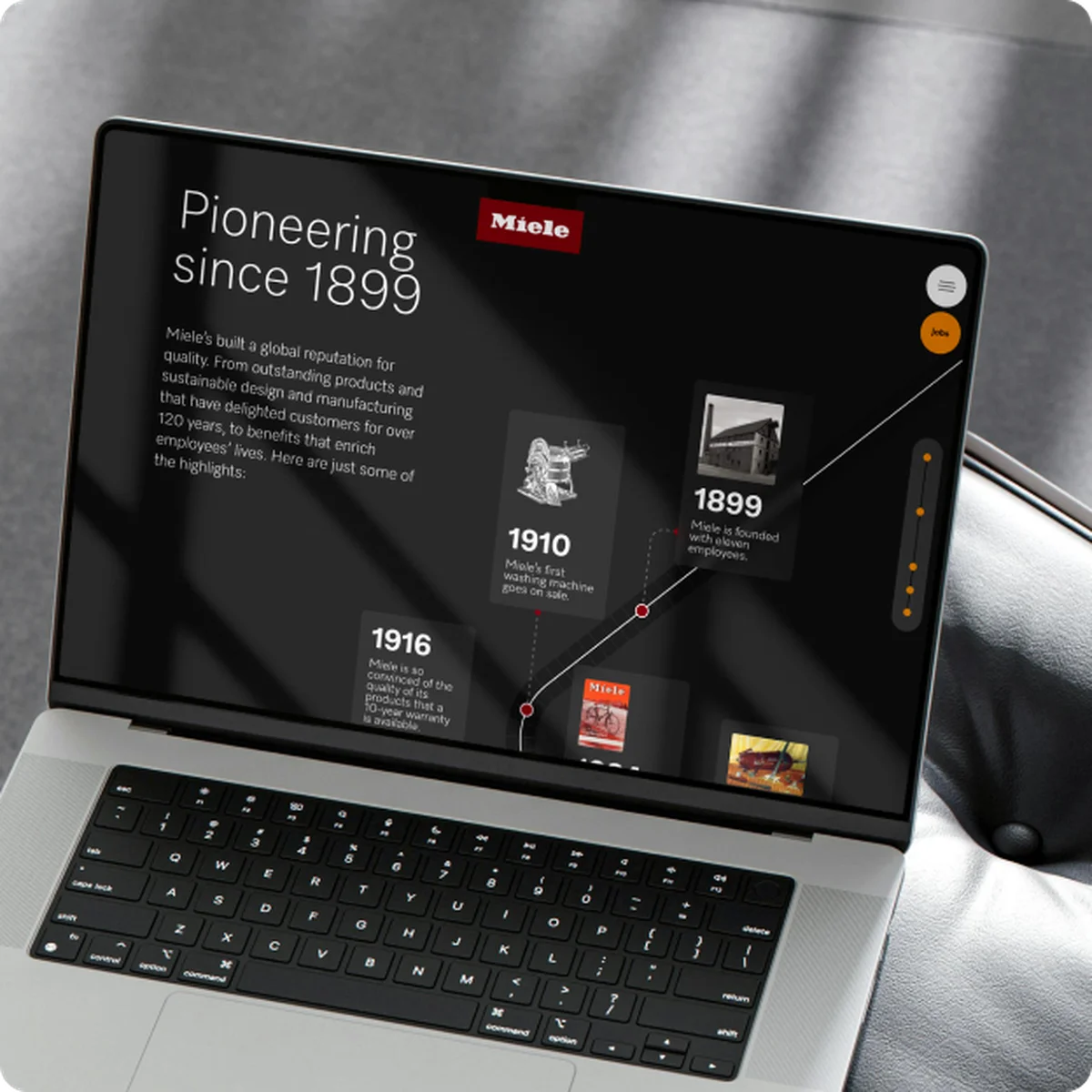 Miele X E-Commerce-Website-Vorschau auf einem Laptop mit einer Markenhistorie-Zeitleiste „Pioneering since 1899“, die Meilensteine wie die erste Waschmaschine von 1910 zeigt, vor einem dunklen Hintergrund in einem hochwertigen Interieur